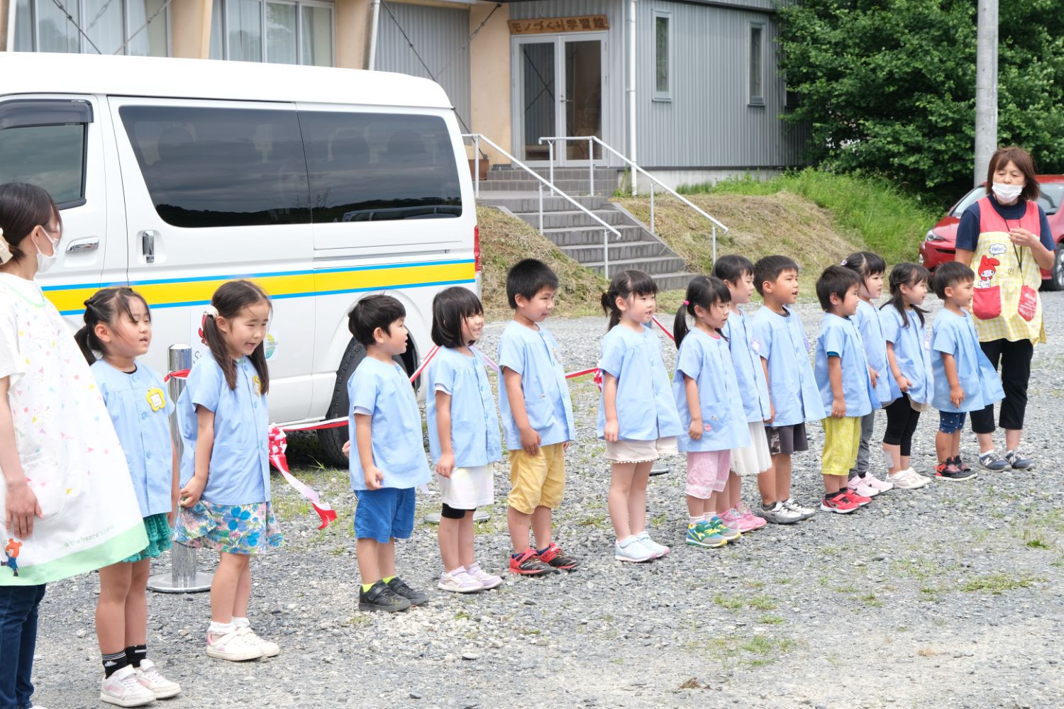 白い送迎バンと階段のある建物の前で、保育士が見守る中、青いスモックを着た幼い子どもたちが並んで立ち、歌をうたっている写真