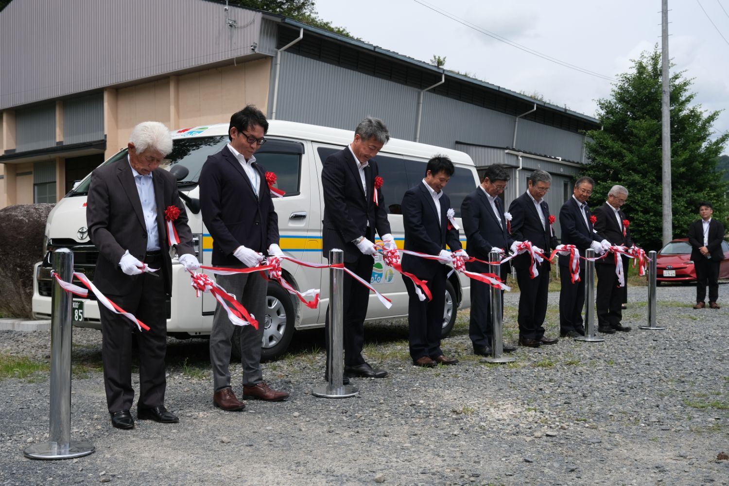 白い送迎バンの前でスーツ姿の男性たちが白手袋をはめて赤白のテープを持ち、一列に並んでテープカットの準備をしている式典の一場面の写真