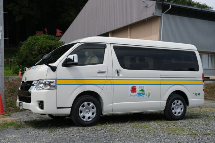 背景には小さな木造建物と斜めの屋根の倉庫がある砂利の駐車場に側面に青と黄色のラインと「Smart GoTo」などのロゴがある白い送迎バンが停まっていている写真