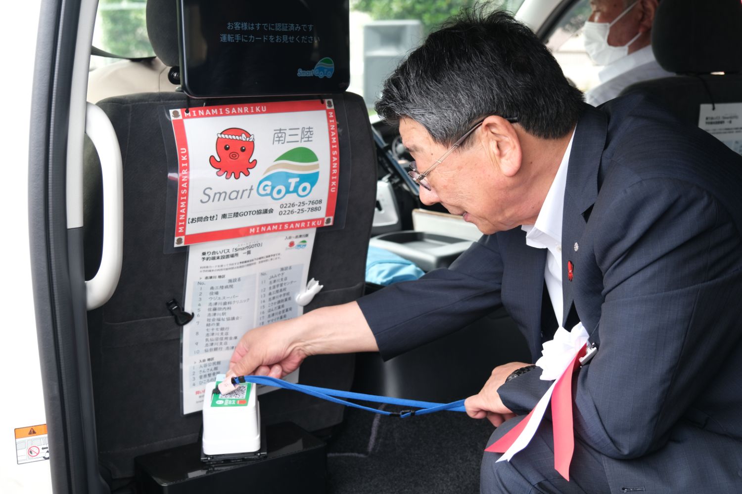 スーツ姿の男性が送迎バンの後部座席のシートに「南三陸 Smart GoTo」と書かれた赤いタコのマスコット付きの案内板の下に設置されたICカード読み取り機に青いストラップ付きのカードをかざしている写真
