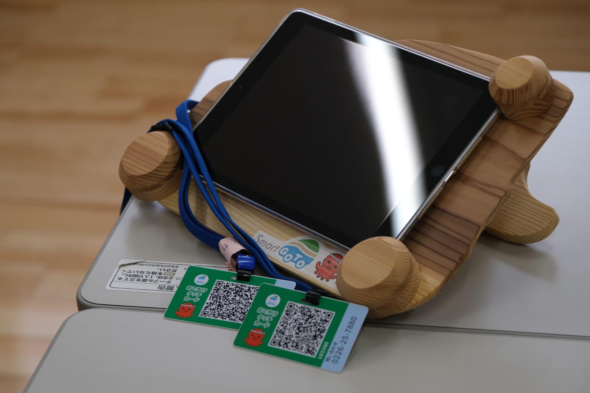 木製の台にタブレット端末が置かれており、その前には「のりかえタッチカード」と書かれたQRコード付きの緑色のカードが2枚、青いストラップに付けられて机の上に並べられている写真
