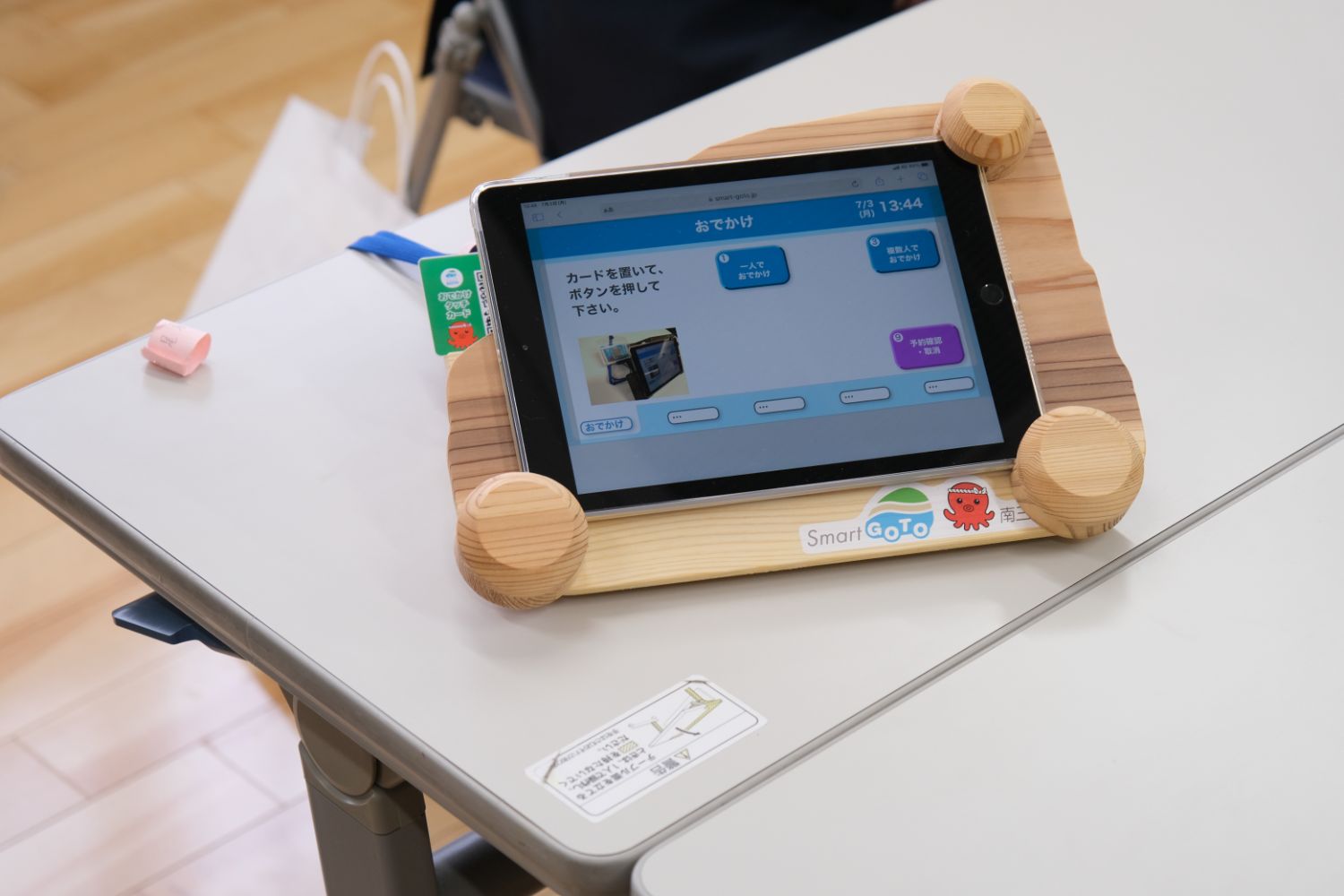 木製の台にセットされたタブレット端末が机の上に置かれており、画面には「カードを置いて、ボタンを押してください」と表示されていて、タブレットの横には緑色のカードが見えている写真