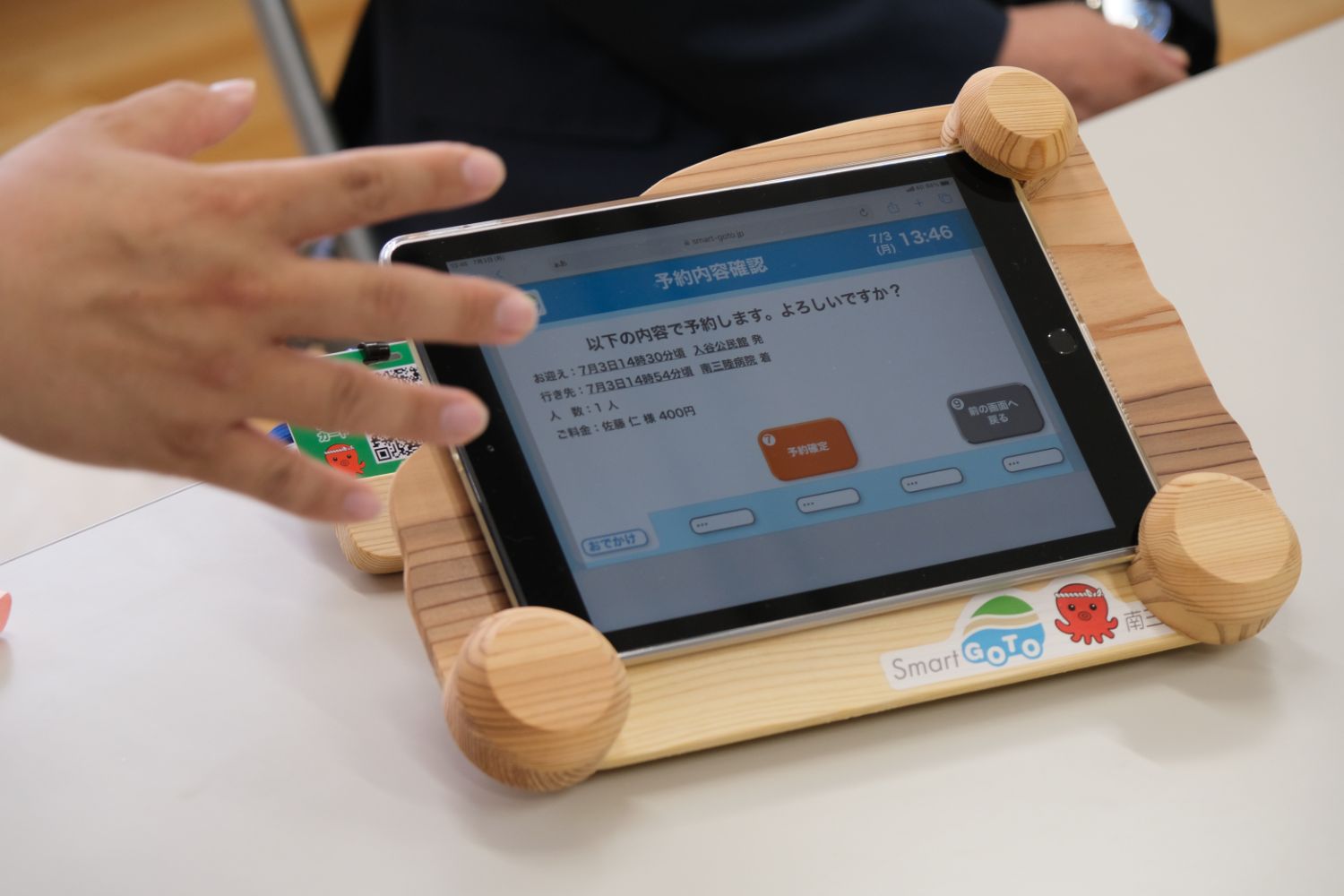タブレットが木製のスタンドに置かれており、画面には「予約内容確認」と表示されたメニューが映っていて、手前には人の手が写っている写真
