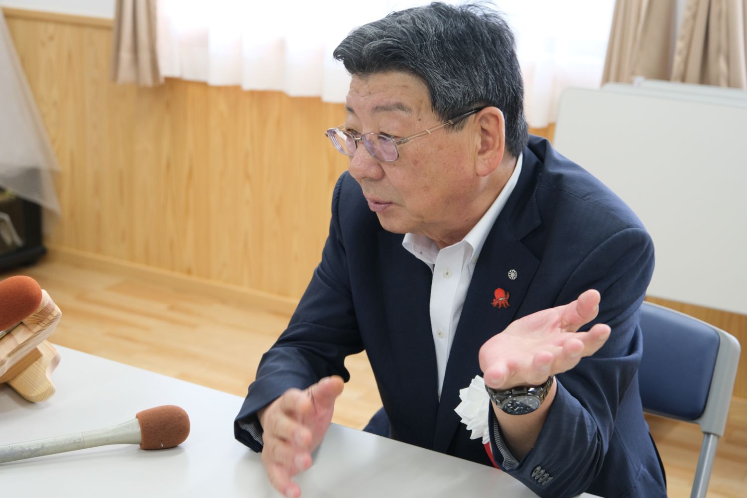 木製の壁とカーテンがかかった窓が見える室内で黒いジャケットを着た男性が机に座り、手を広げて話している写真