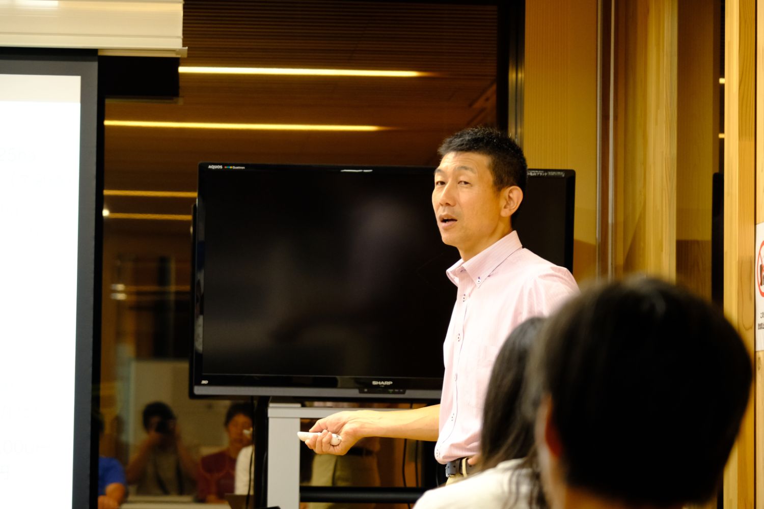 ピンクのシャツを着た講師がスクリーンの前で話をしており、後方には真剣に聞き入る受講者たちが映っている、ブルーカーボン研修の講義中の写真