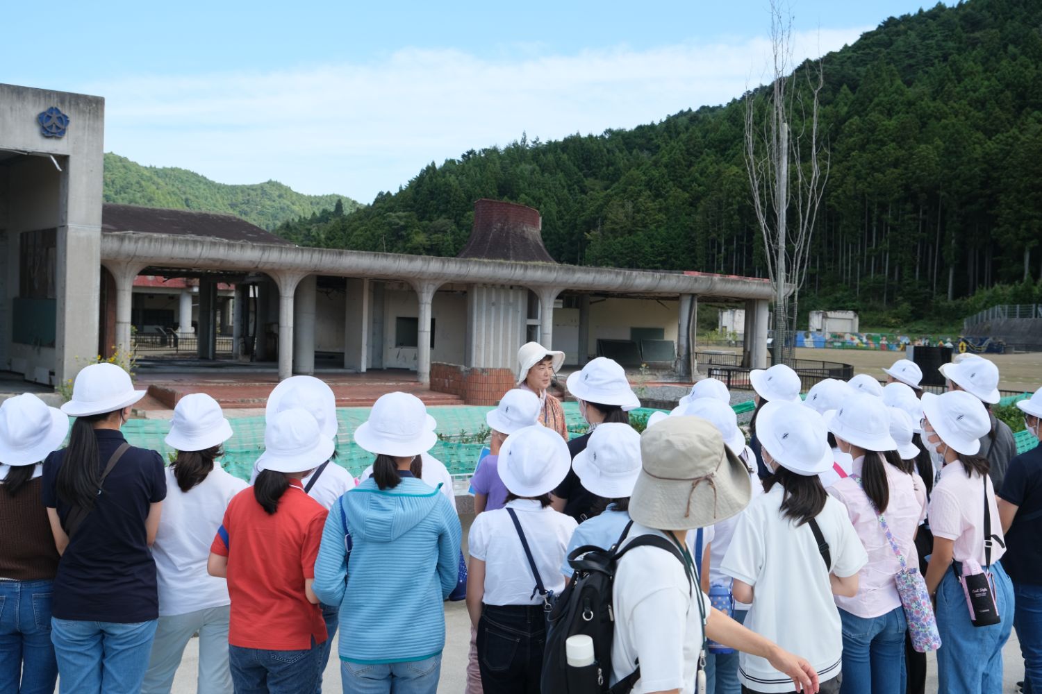 背景には緑の山々と古い建物があり、白い帽子をかぶった子どもたちが屋外で先生の話を聞いている様子の写真