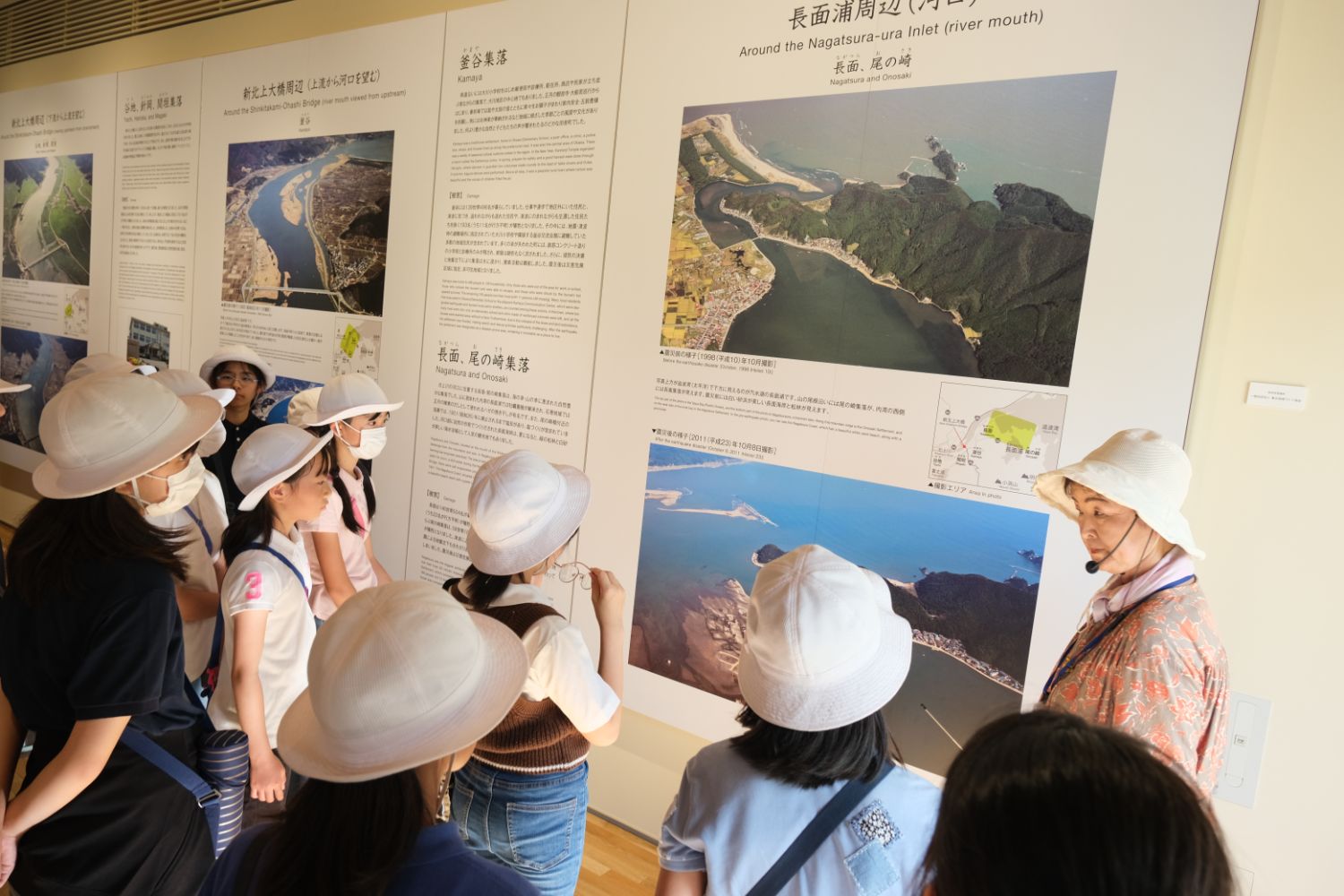 展示パネルの前で、白い帽子をかぶった子どもたちが、説明員の女性の話を聞きながら地形や地域に関する写真や解説を見て学んでいる様子の写真