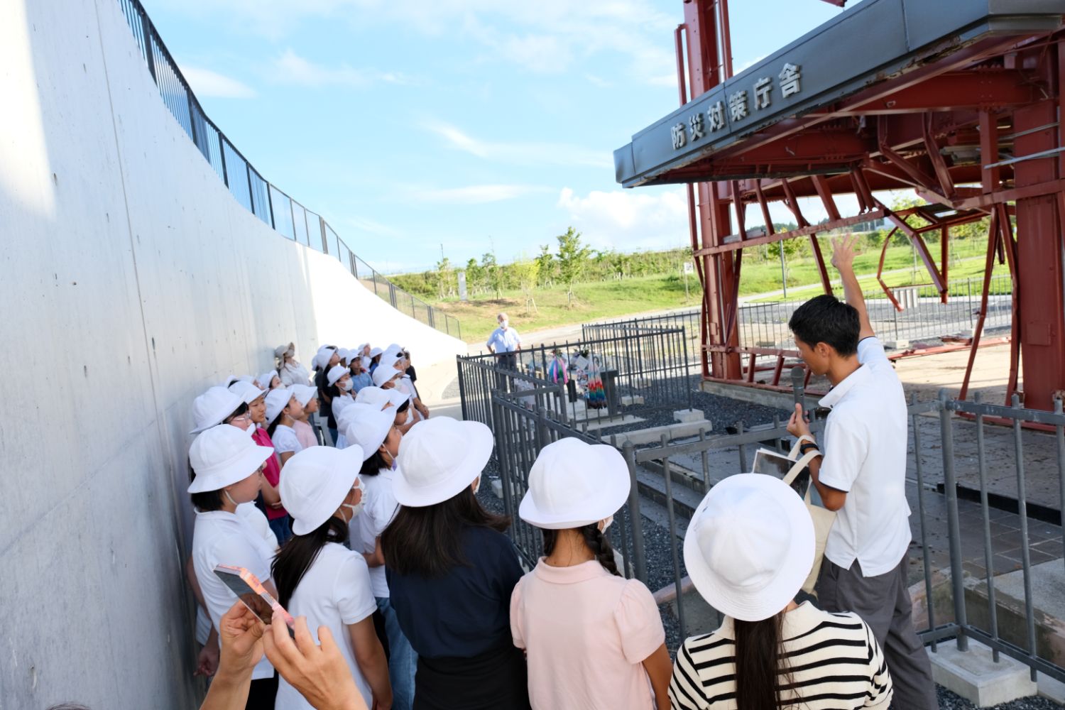 男性が津波の被害を受けた鉄骨の建物の一部を指さしながら説明しており、白い帽子をかぶった子どもたちのグループが真剣に聞いている写真