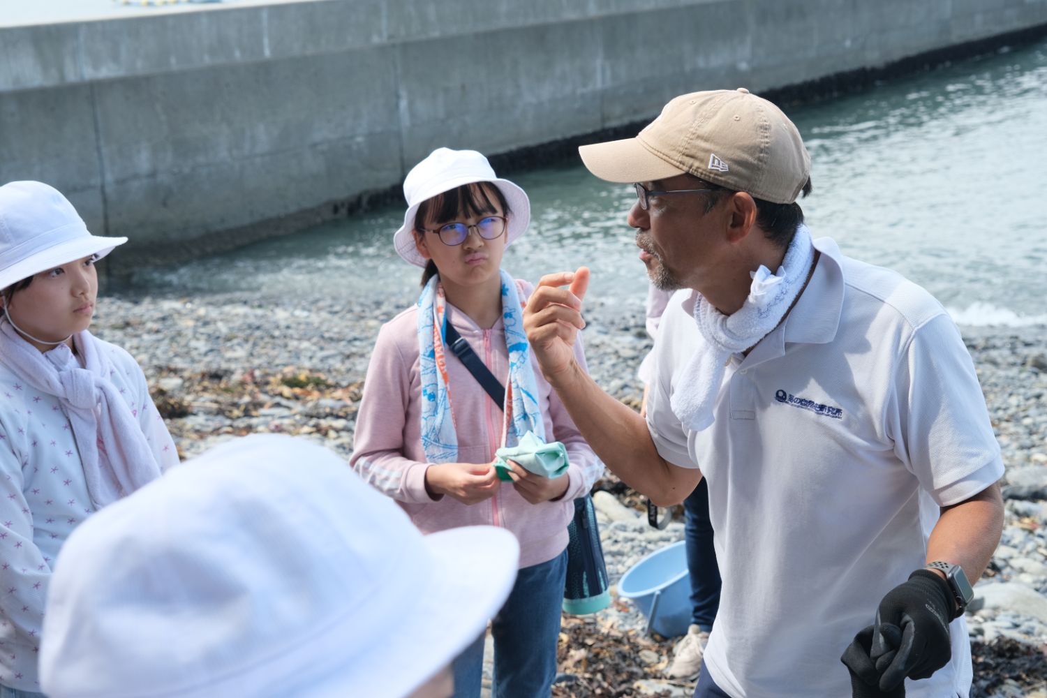海岸の石浜で、帽子をかぶった子どもたちが男性の説明を真剣に聞いている写真