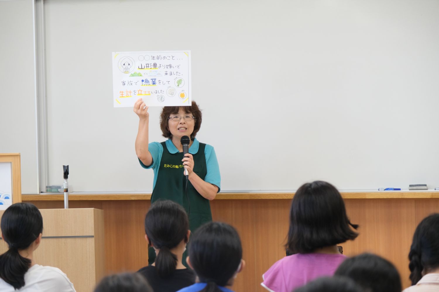 教室で民子さんがマイクを持ち、手作りの紙を掲げて子どもたちに話している様子の写真