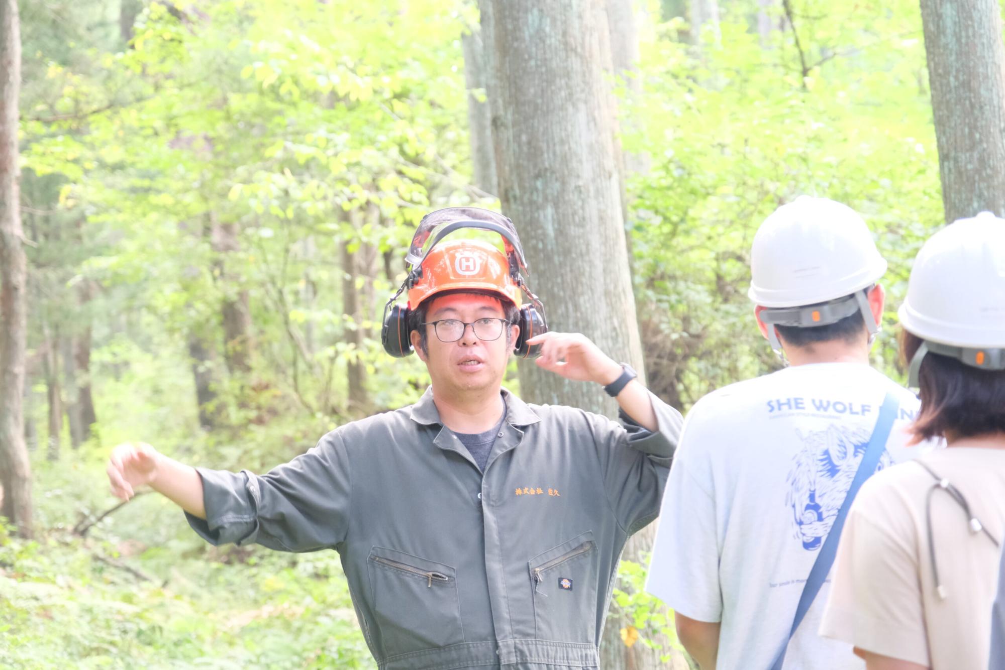 森林の中で佐藤太一さんがオレンジの防護ヘルメットを着用し、身振りを交えて林業体験の説明をしている様子の写真