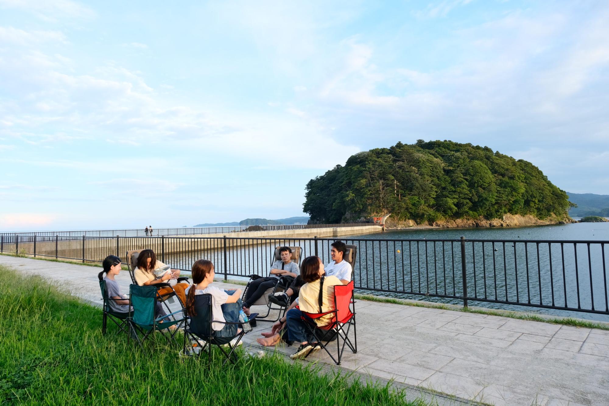 海辺の遊歩道で参加者たちが円形に椅子を並べて座り、海と小島を背景に穏やかな時間を共有している様子の写真