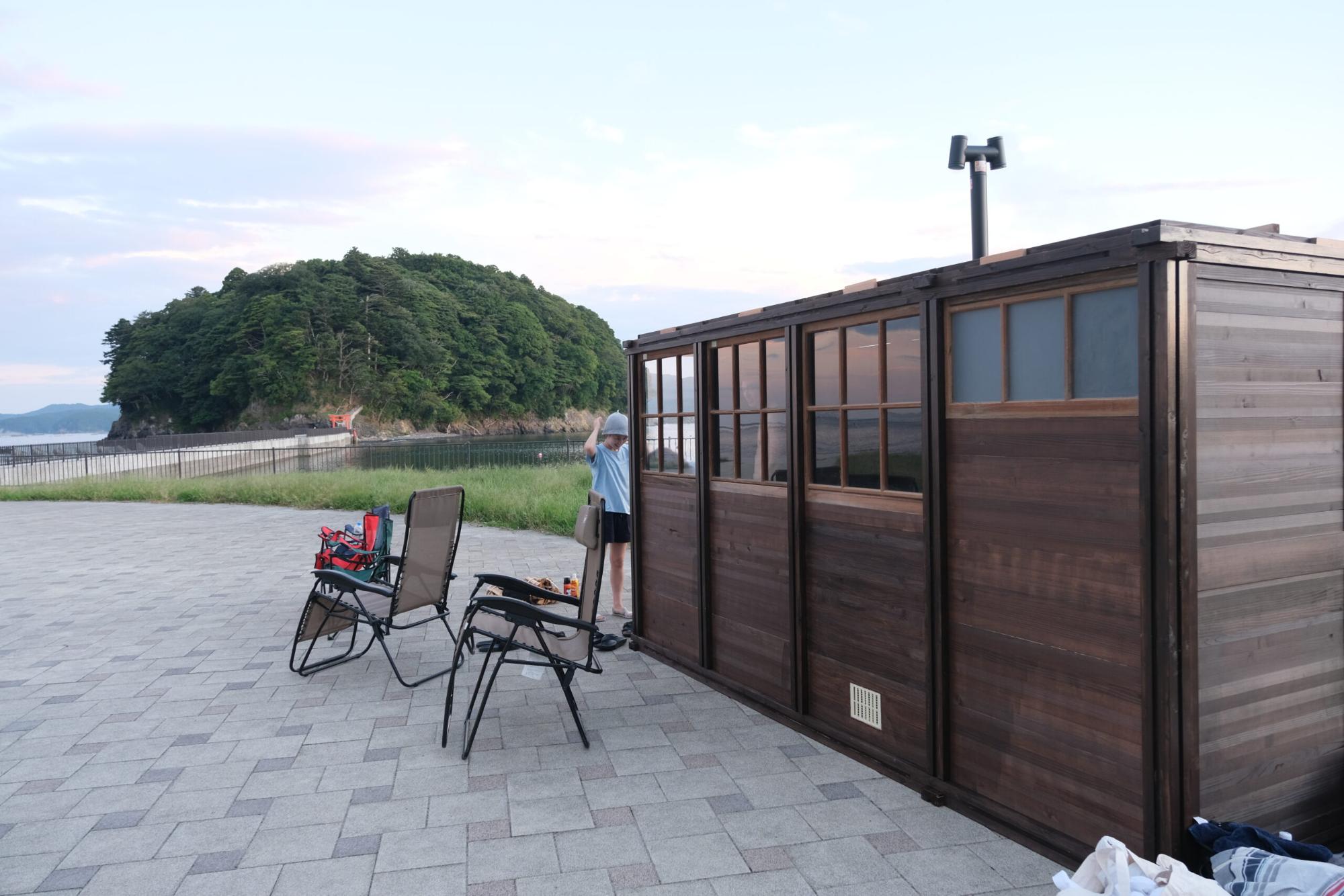 海辺の広場に設置された木製のサウナ小屋の前で、帽子をかぶった人が準備をしており、近くにはリクライニングチェアと荷物が置かれている写真