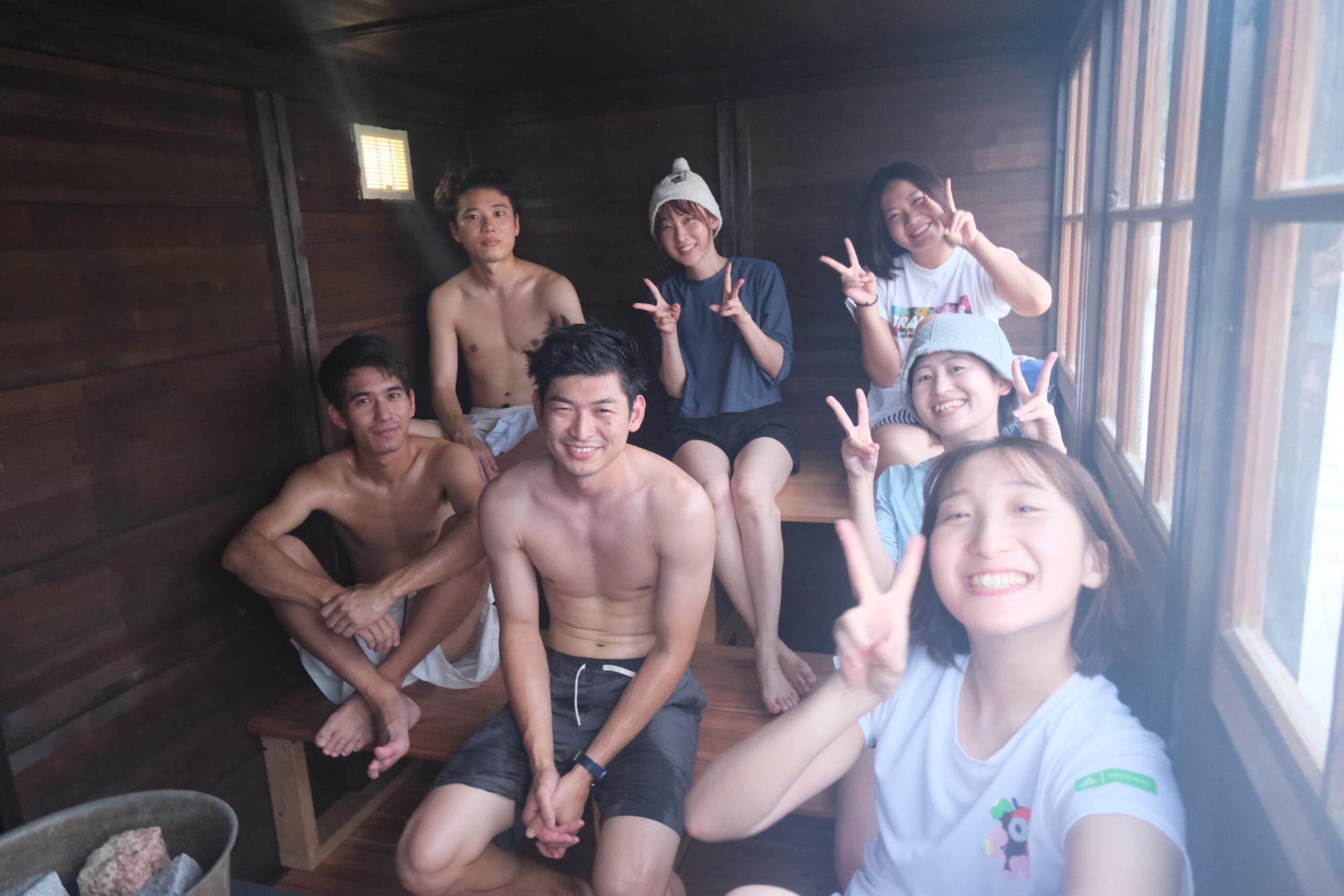 木の内装のサウナ室の中で、若い男女のグループがリラックスしながら笑顔でピースサインをしている海辺のサウナ体験の写真