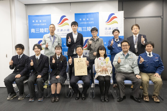 南三陸町と志津川湾のロゴが入った背景の前で、賞状を持った町長と高校生、関係者の方々を含む12人が並んで座り、全員が親指を立て「サムズアップ」のポーズをして写っている集合写真