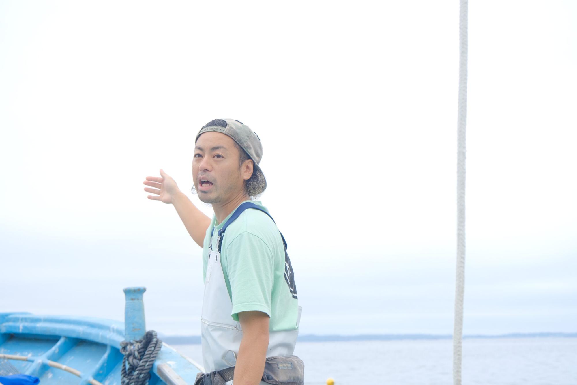 海上の船の上で、帽子を後ろ向きにかぶった阿部和也さんが腕を伸ばして沖の方向を指差しながら、海について説明している写真
