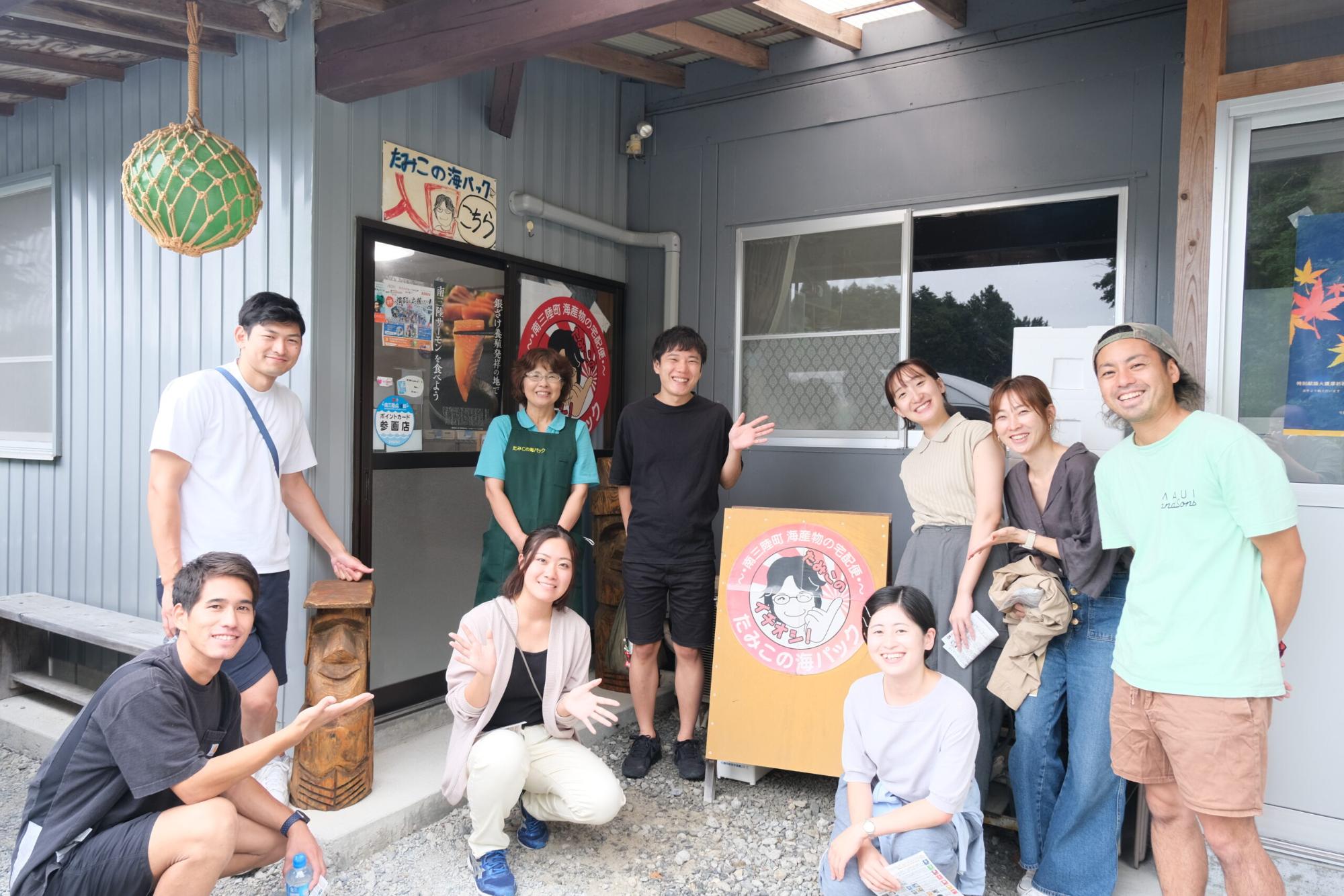 地域体験施設の入口前で「たみこの海パック」と書かれた看板の前に、案内人の女性と若者たちが笑顔で並び、両手を広げてポーズを取っている集合写真