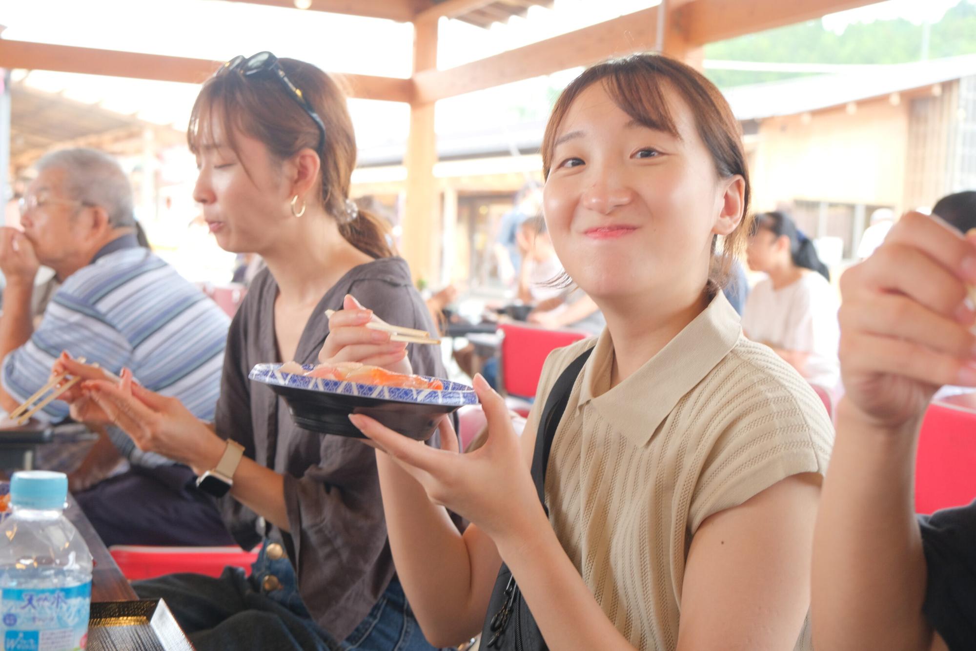 屋根付きの食事スペースで、女性が海鮮丼を手に笑顔で口に運んでおり、周囲では他の参加者たちも同様に食事を楽しんでいる地域の食体験の写真