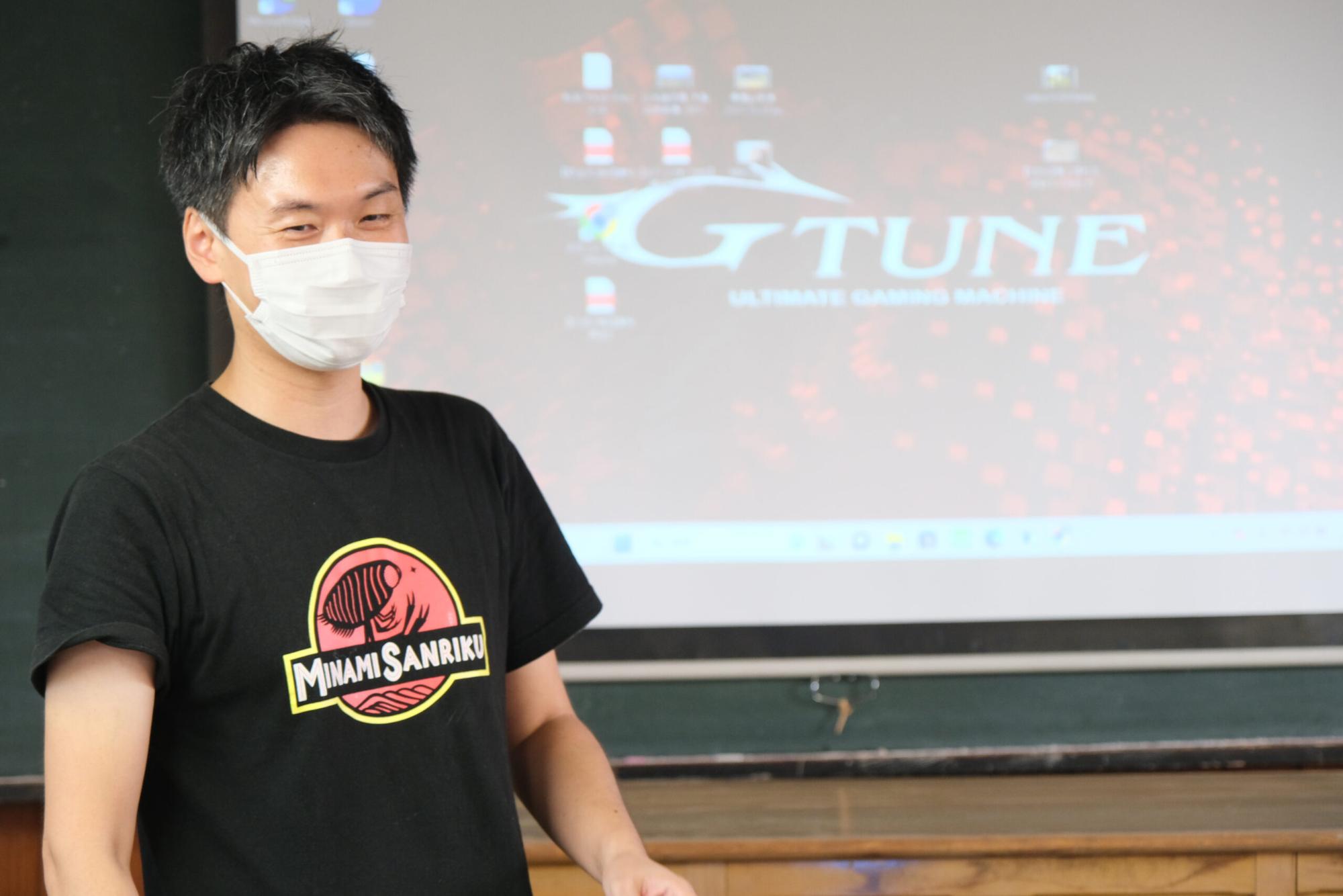 黒いTシャツに「MINAMI SANRIKU」と書かれたロゴが入った服を着た男性スタッフが、マスク姿で教室の前に立ち、パソコン画面をバックに話をしている写真