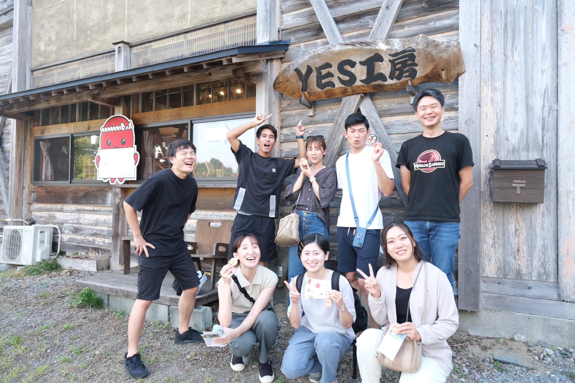 木造の建物「YES工房」の前で、若者たちが笑顔でポーズを取り、それぞれが体験で作った作品やパンフレットを手に持ち体験工房の集合写真