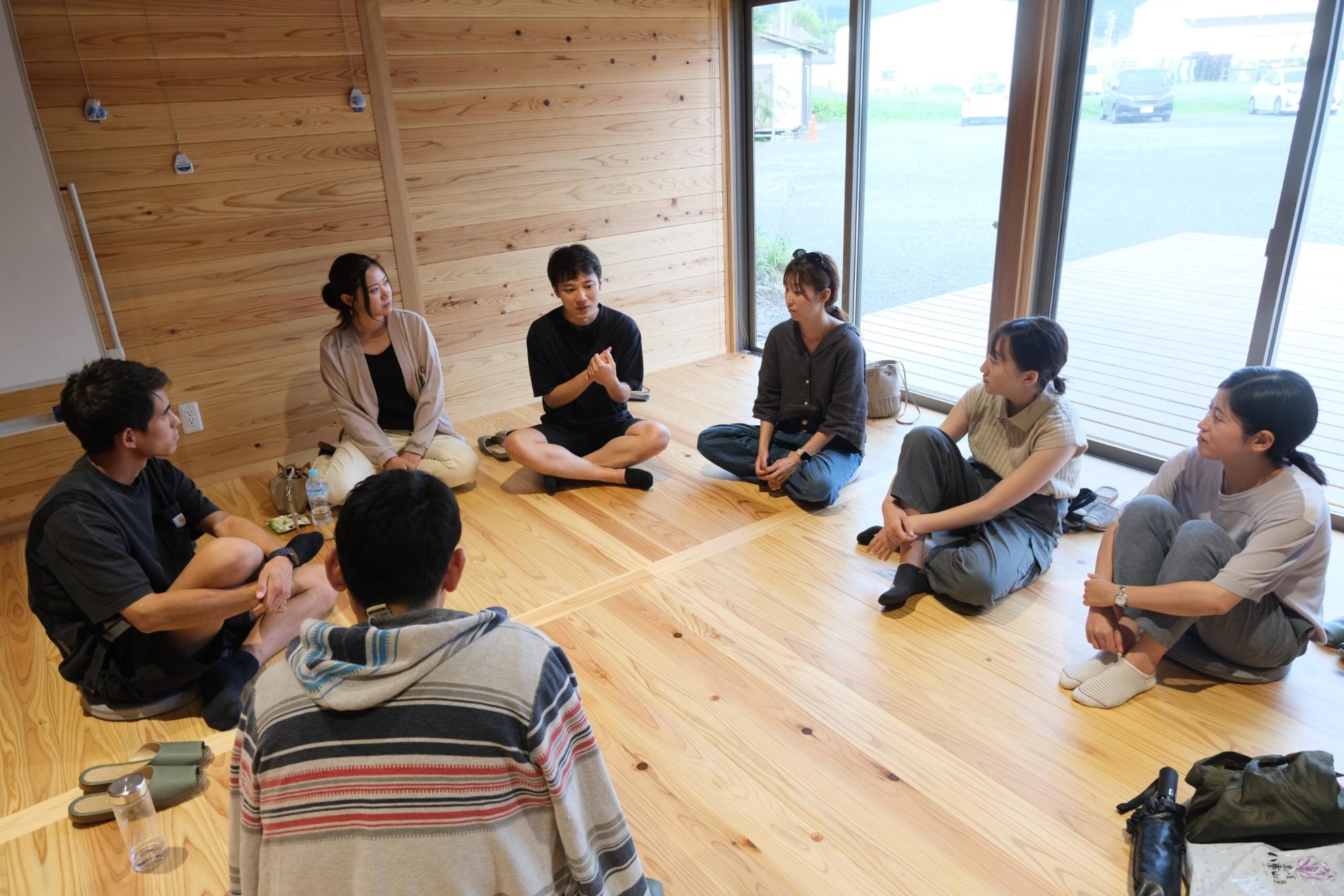 木造の室内で参加者たちが円になって座り、一人の男性が手振りを交えて話すのを他のメンバーが集中して聞いている様子の写真