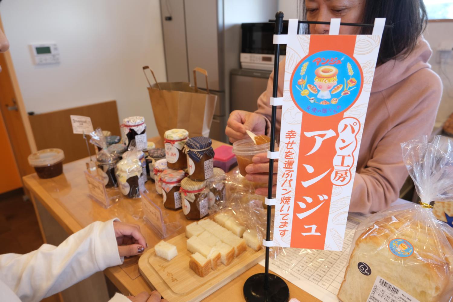 ジャムや食パンが並ぶ販売コーナーで、女性が透明カップからスプーンですくってパンに塗るジャムを試食している様子の写真