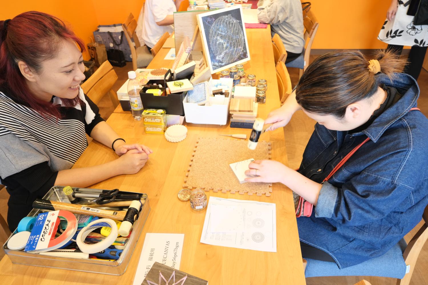 テーブルの上に並べられた道具を使い、女性が木板に釘を打ちながらハンドメイド作品を制作し、もう一人が笑顔で見守っている様子の写真