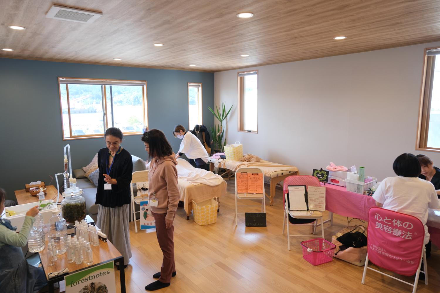 木の床と明るい窓がある室内で、マッサージやアロマ体験、健康相談など複数のブースが並び、来場者とスタッフが会話したり施術を受けたりしている様子の写真