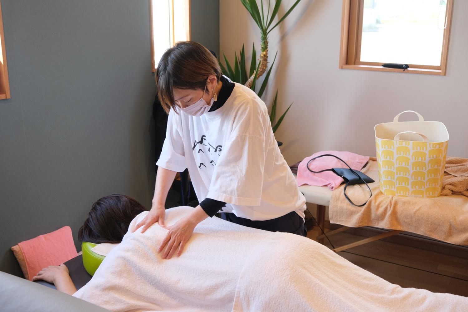 ベッドにうつ伏せになった人の背中を、白いTシャツを着たスタッフが両手で丁寧にマッサージしている様子の写真