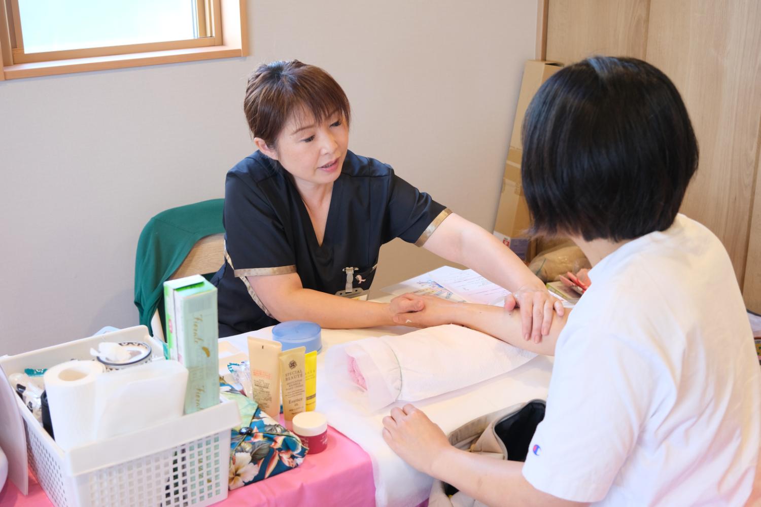 施術スタッフがテーブル越しに座る来場者の腕を優しくさすりながらスキンケアを行っており、机の上にはクリームやローションなどのケア用品が並んでいる、ハンドケア体験の写真