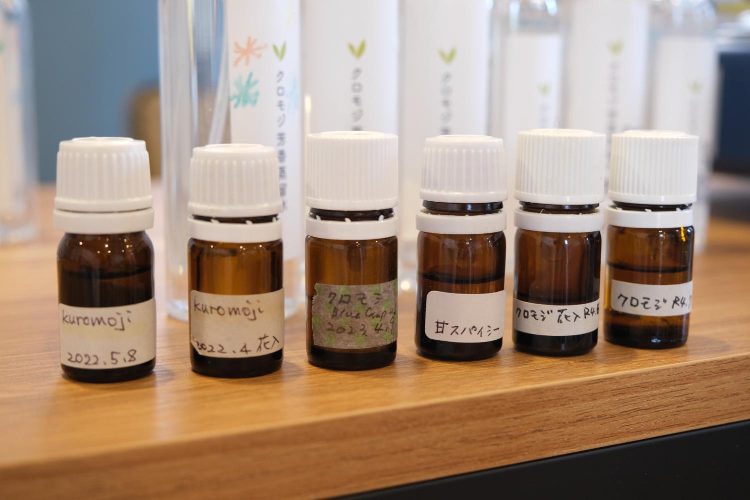 ラベルに「クロモジ」や「甘スパイシー」などと手書きされた小さな茶色の瓶が6本並び、後方にはスプレーボトルも見える、アロマオイルの展示の写真