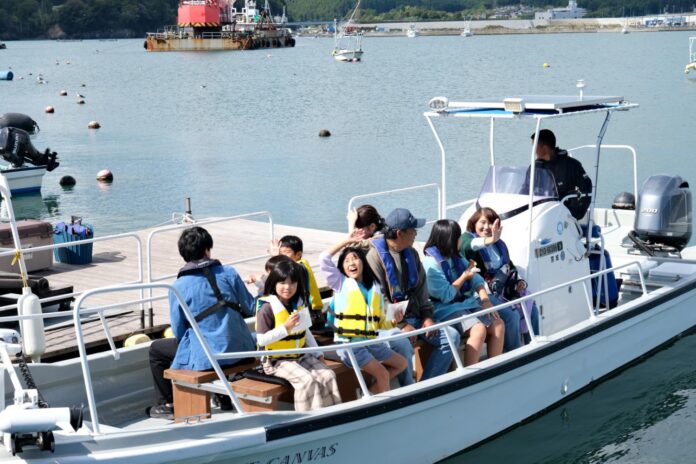 子どもと大人のグループがライフジャケットを着て桟橋に停泊した小型ボートに乗り込み、笑顔で手を振りながら出発を待っている様子の写真