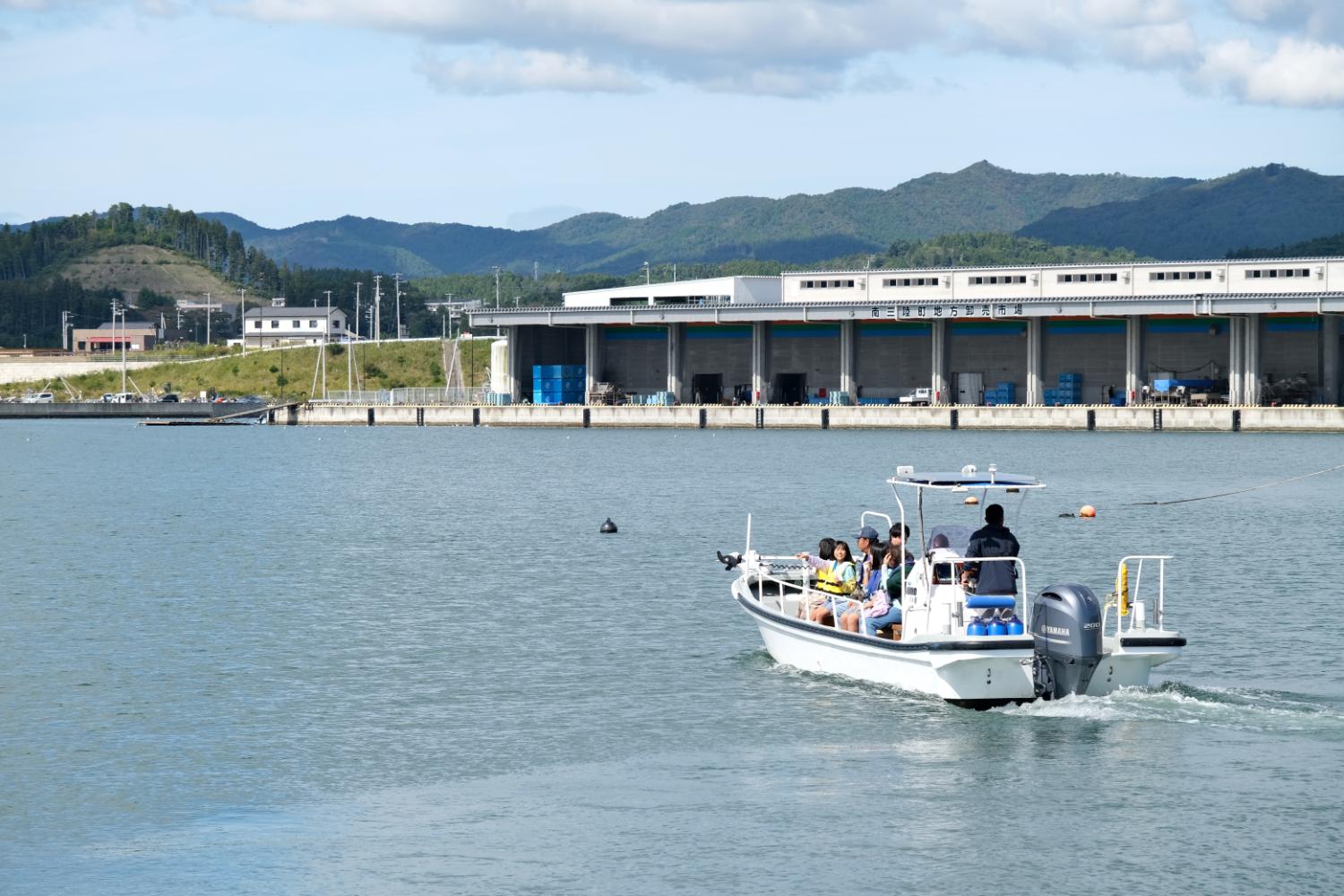 ライフジャケットを着た複数の人たちが港から出発する小型ボートに乗っており、穏やかな海と背景にある建物が見える、港での乗船体験の写真