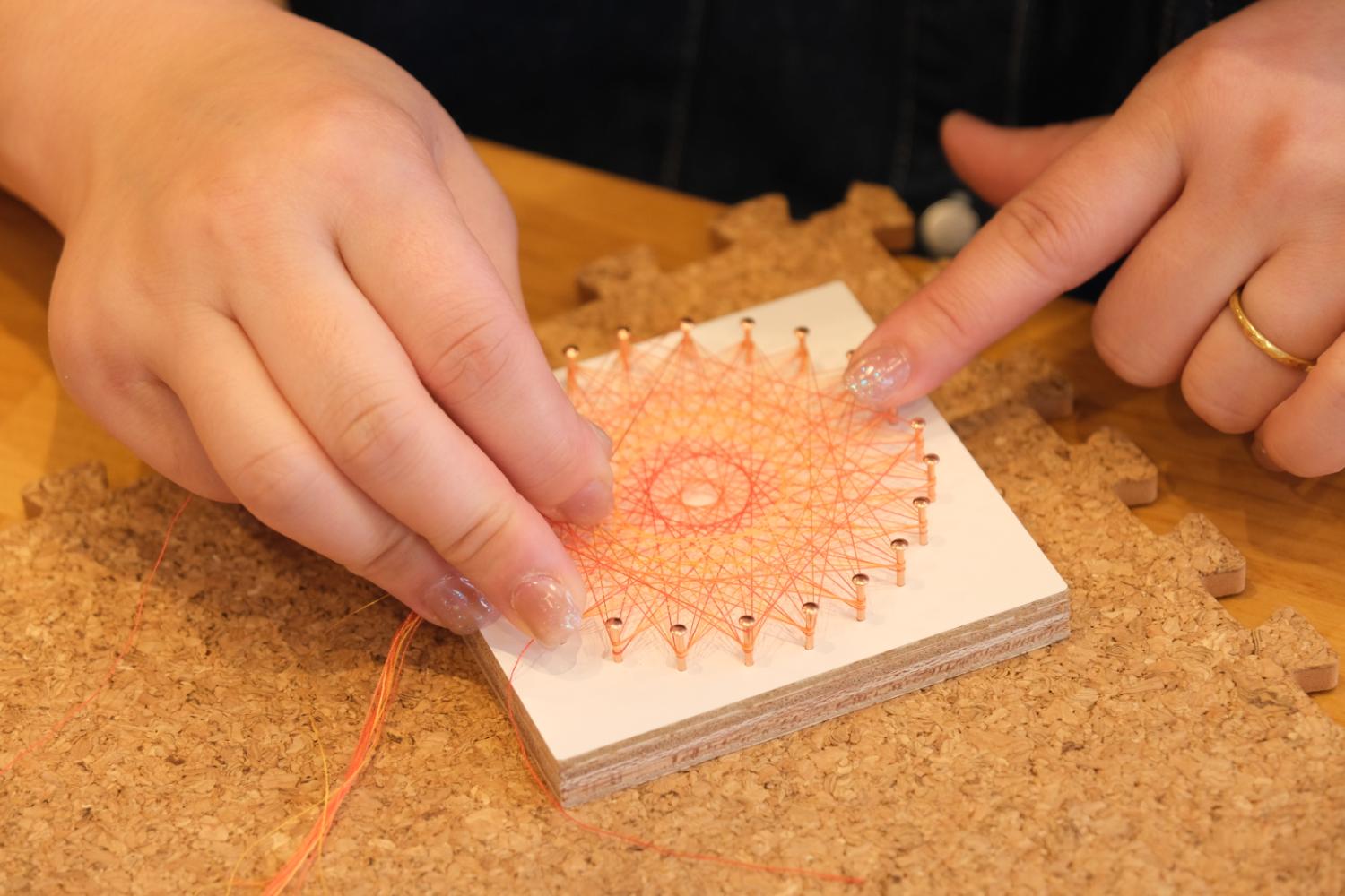 木の板に打たれた円形の釘にオレンジ色の糸を何重にもかけて模様を作る手元の様子を写した、糸かけアート体験の写真