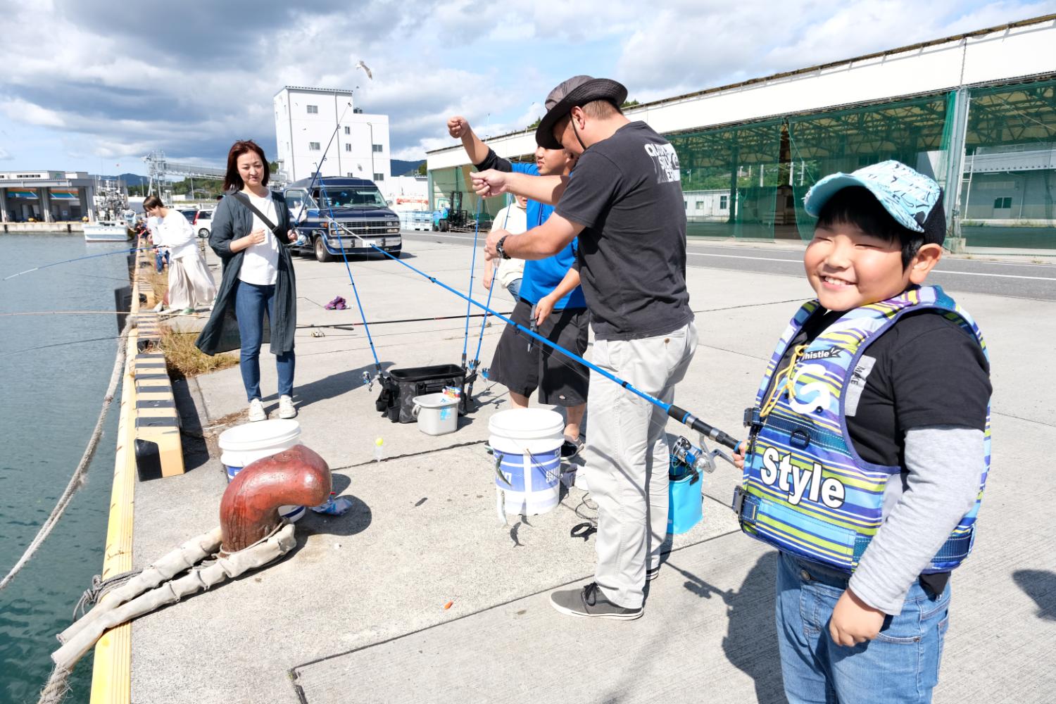 青い空の下、港の岸壁で釣り竿を持った大人たちが釣りを楽しんでおり、手前ではライフジャケットを着た男の子が笑顔でカメラを見ている、親子釣り体験の写真