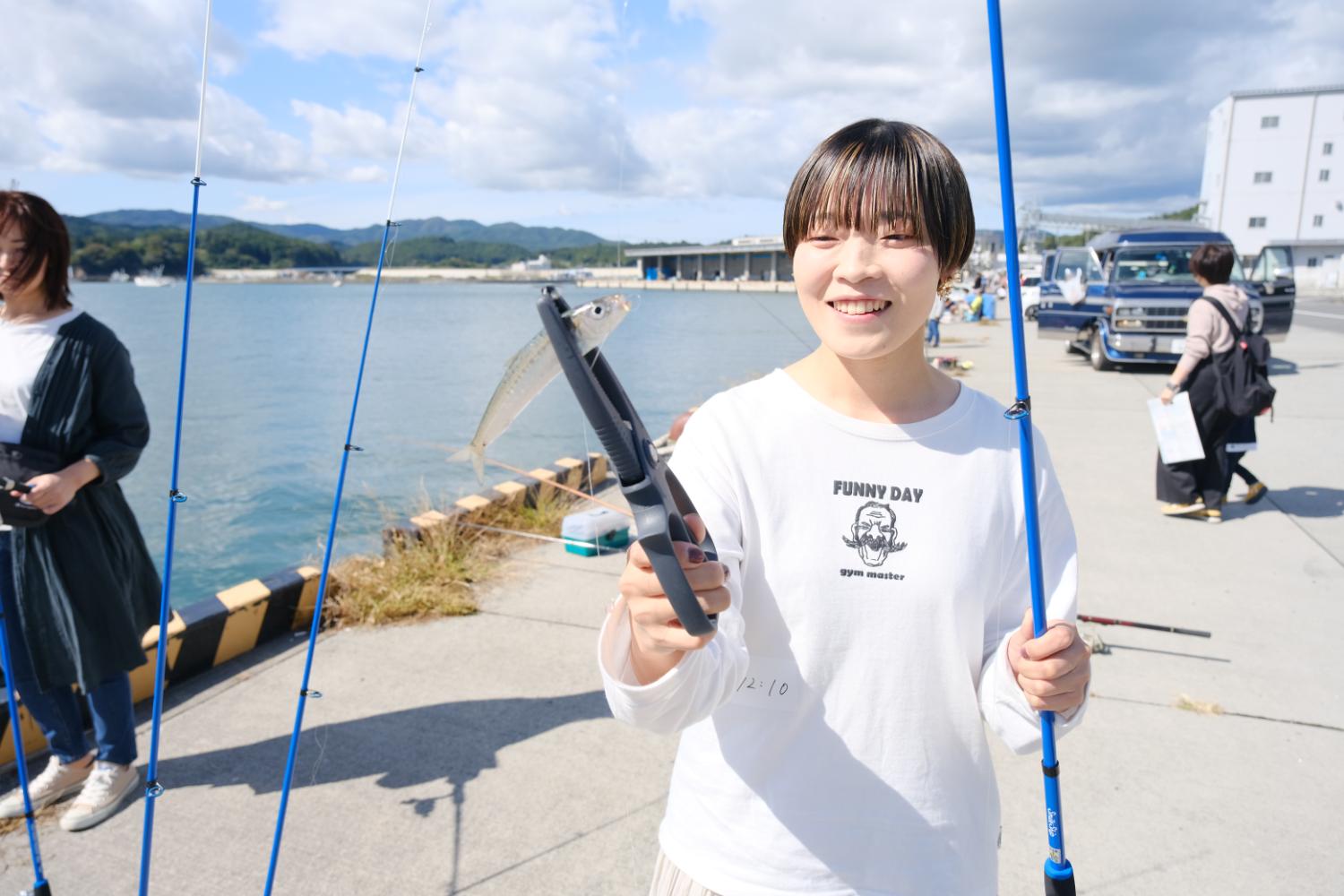 白いシャツを着た女性が釣り竿を片手に、もう一方の手でトングのような道具を使って釣れた魚を笑顔で掲げている写真
