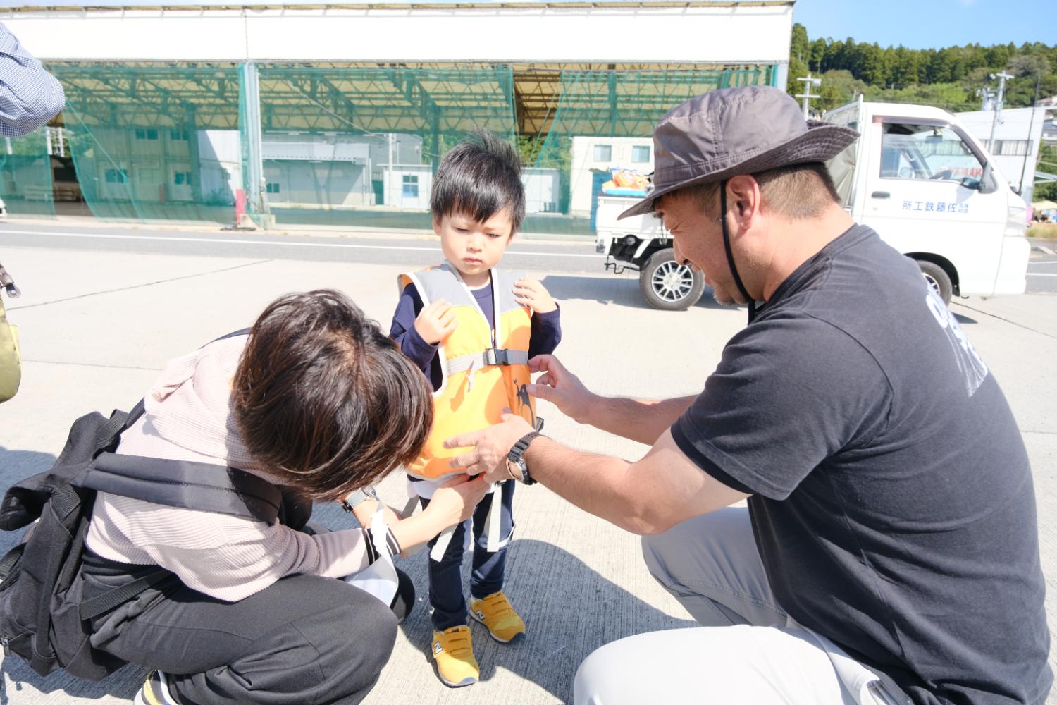 港の岸辺で男の子に大人二人がオレンジ色のライフジャケットを丁寧に着せている写真