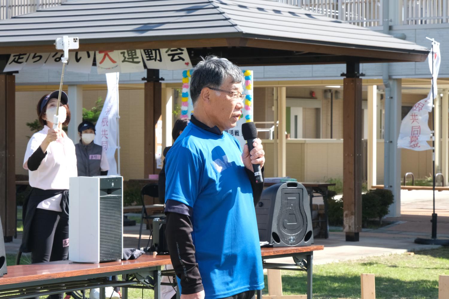 背景には大会名の垂れ幕や運営スタッフがおり、青いTシャツを着た鈴木さんがマイクを持って開会式のあいさつをしている写真