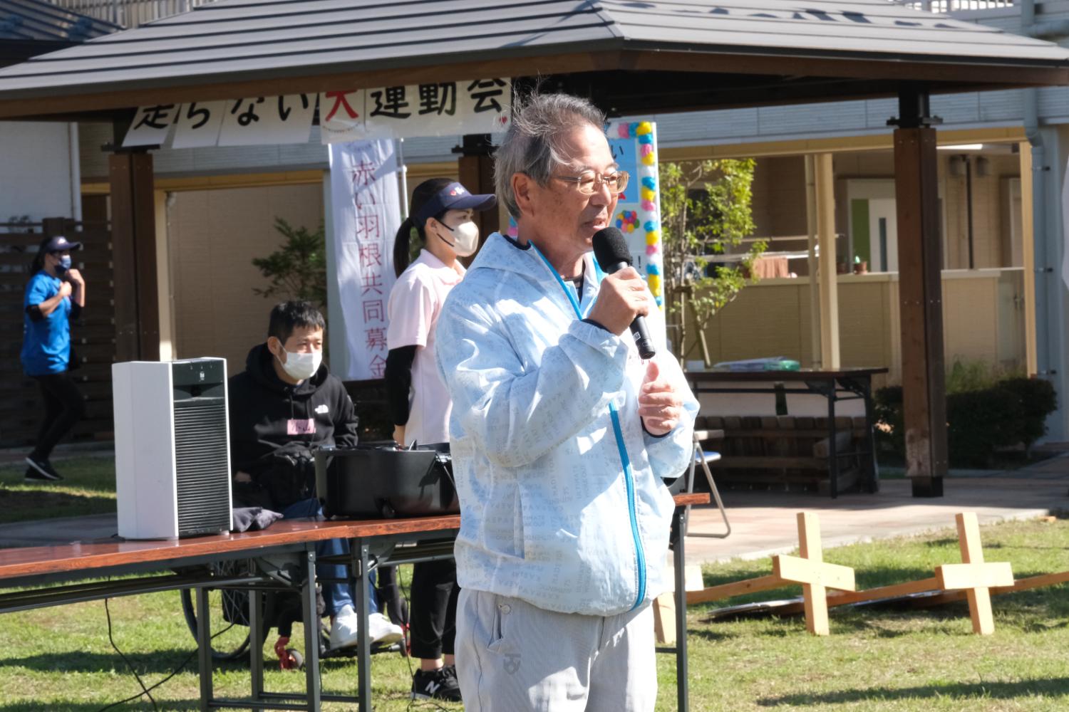 周囲にはスタッフや参加者が立ち、白いジャンパーを着た佐藤会長がマイクを持って挨拶をしている写真