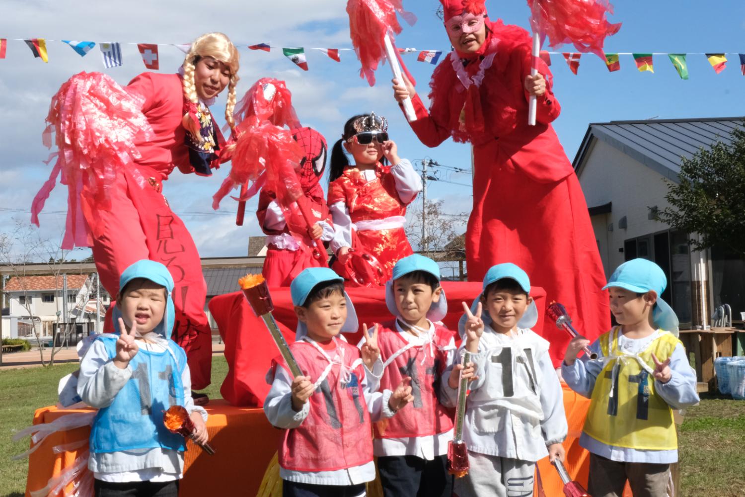赤い衣装を着た大人が高台の上でポンポンを持ち、前列には青い帽子をかぶった幼稚園児たちが色とりどりの衣装を着て並び、ピースサインをしている写真