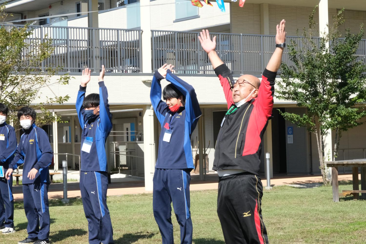 屋外で青いジャージを着た高校生と赤と黒の服を着た男性が、両手を頭の上に上げて元気よく準備体操をしている様子の写真
