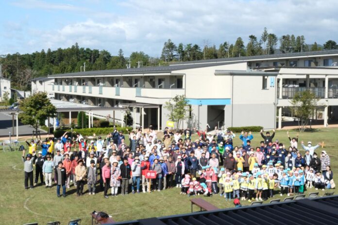 芝生の広場に高齢者、子ども、保育園児、地域住民などが整列して、晴天の下でみんなが手を振っている集合写真