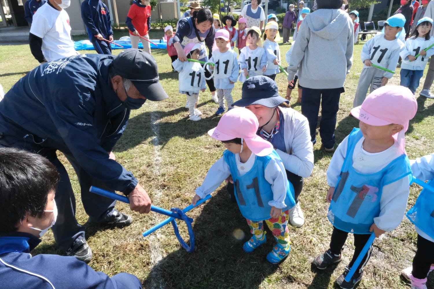 青いビブスとピンクの帽子をかぶった幼児が、大人に手伝ってもらいながら青い輪のついた道具を使う練習をしている様子の写真