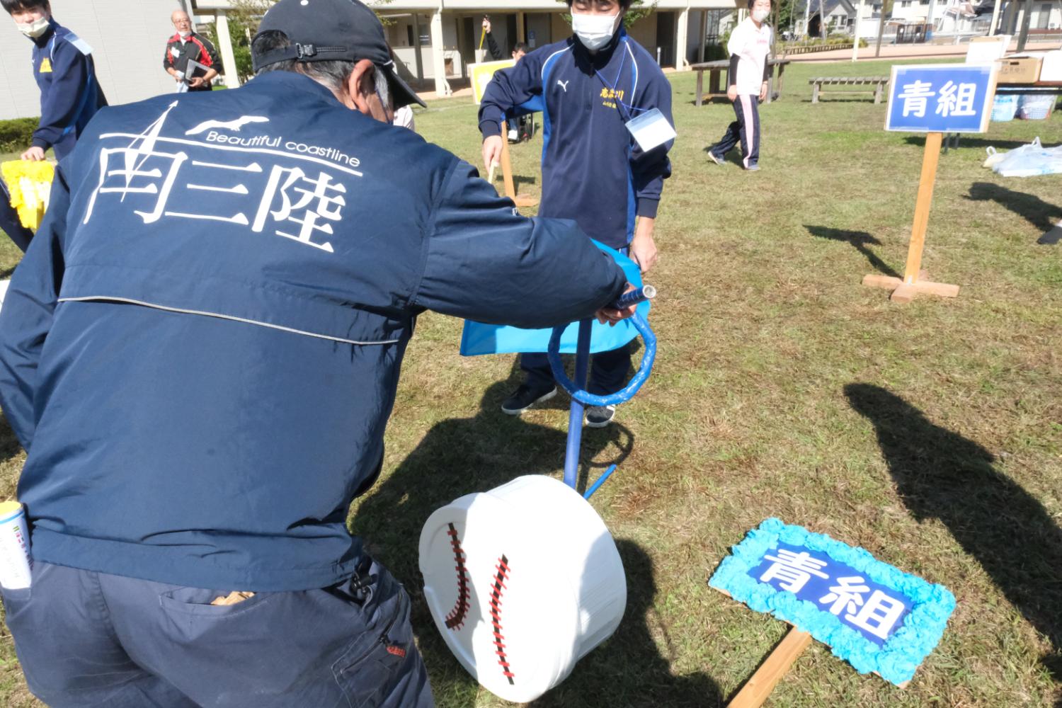 「南三陸」と書かれた上着を着た男性が、青組の道具を調整しながら、背後で高校生と一緒に競技の準備をしている様子の写真