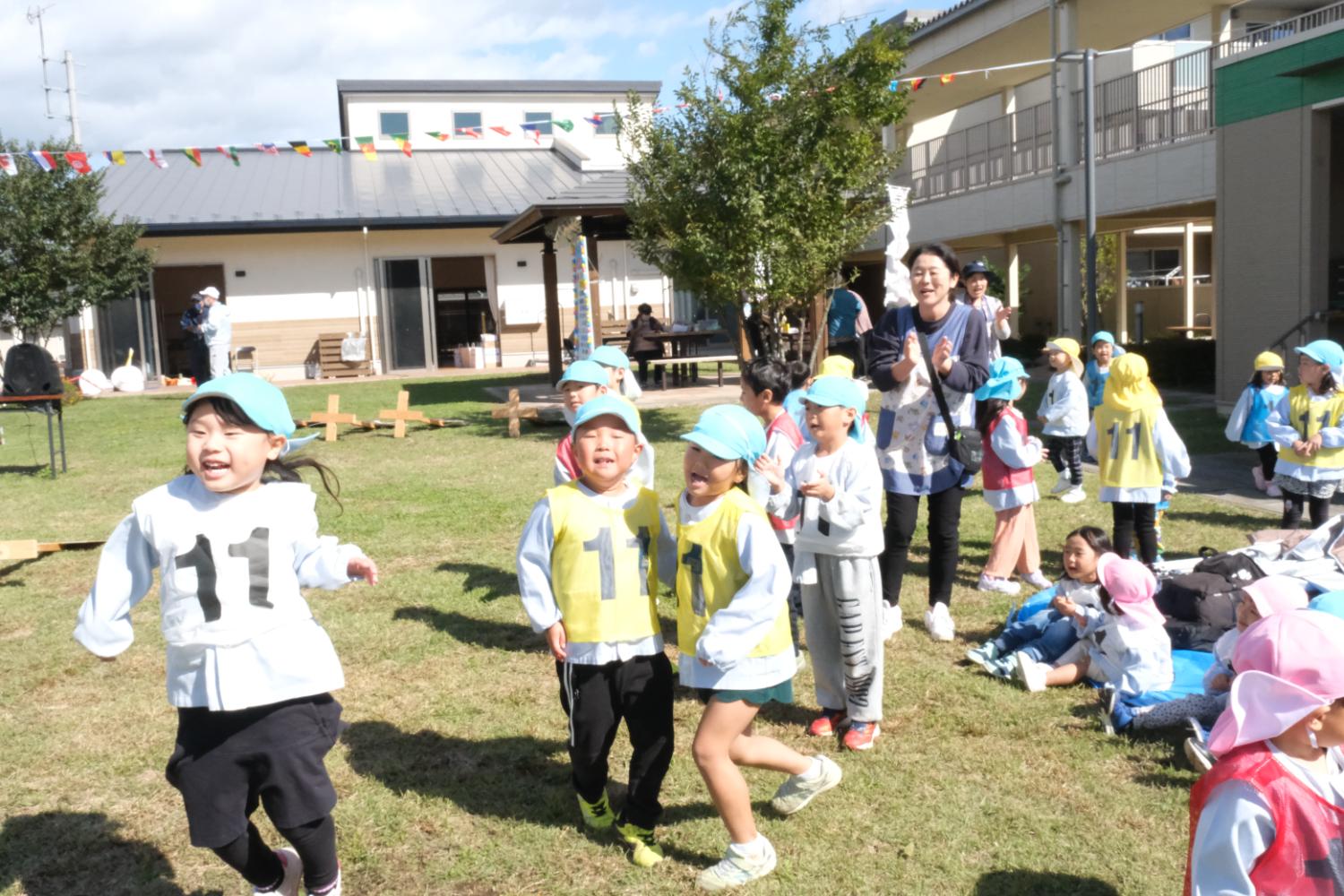 青い帽子をかぶった幼児たちが、番号「11」のビブスを着て芝生の上で元気に走り回り、後ろで大人たちが笑顔で拍手して応援している様子の写真