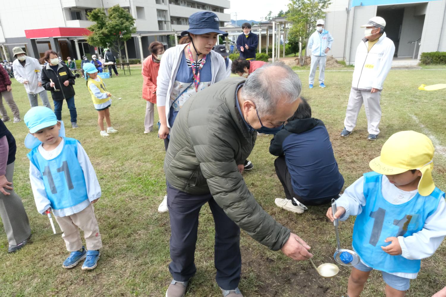 高齢の男性がしゃがんで、黄色の帽子をかぶった幼児にスプーンで水を渡すように手助けしている、スプーンリレーの場面の写真