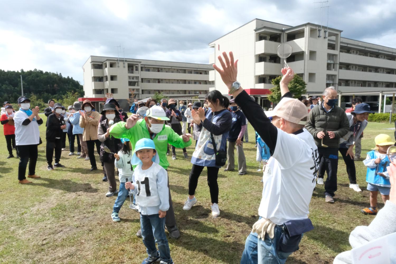 緑色のビブスを着た大人たちと子どもたちがスプーンを持ちながら両手を上げて喜んでおり、後ろでは観客が拍手している様子の写真
