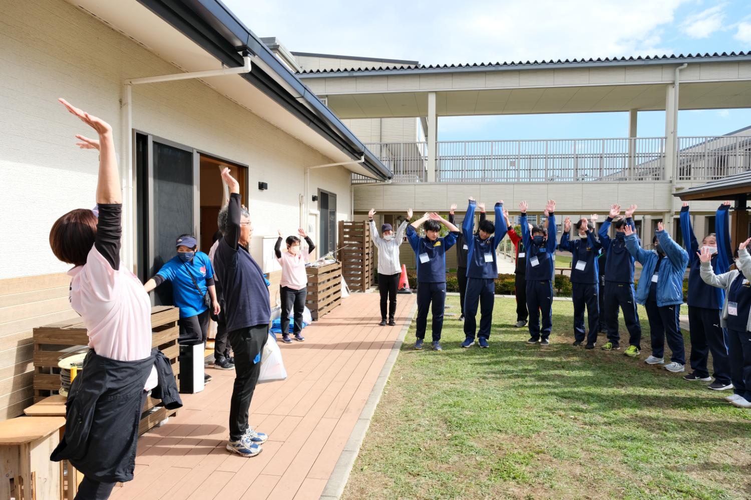 建物の外の通路と芝生の広場で、高校生や大人たちが円をつくって立ち、両手を高く上げて万歳をしている様子の写真