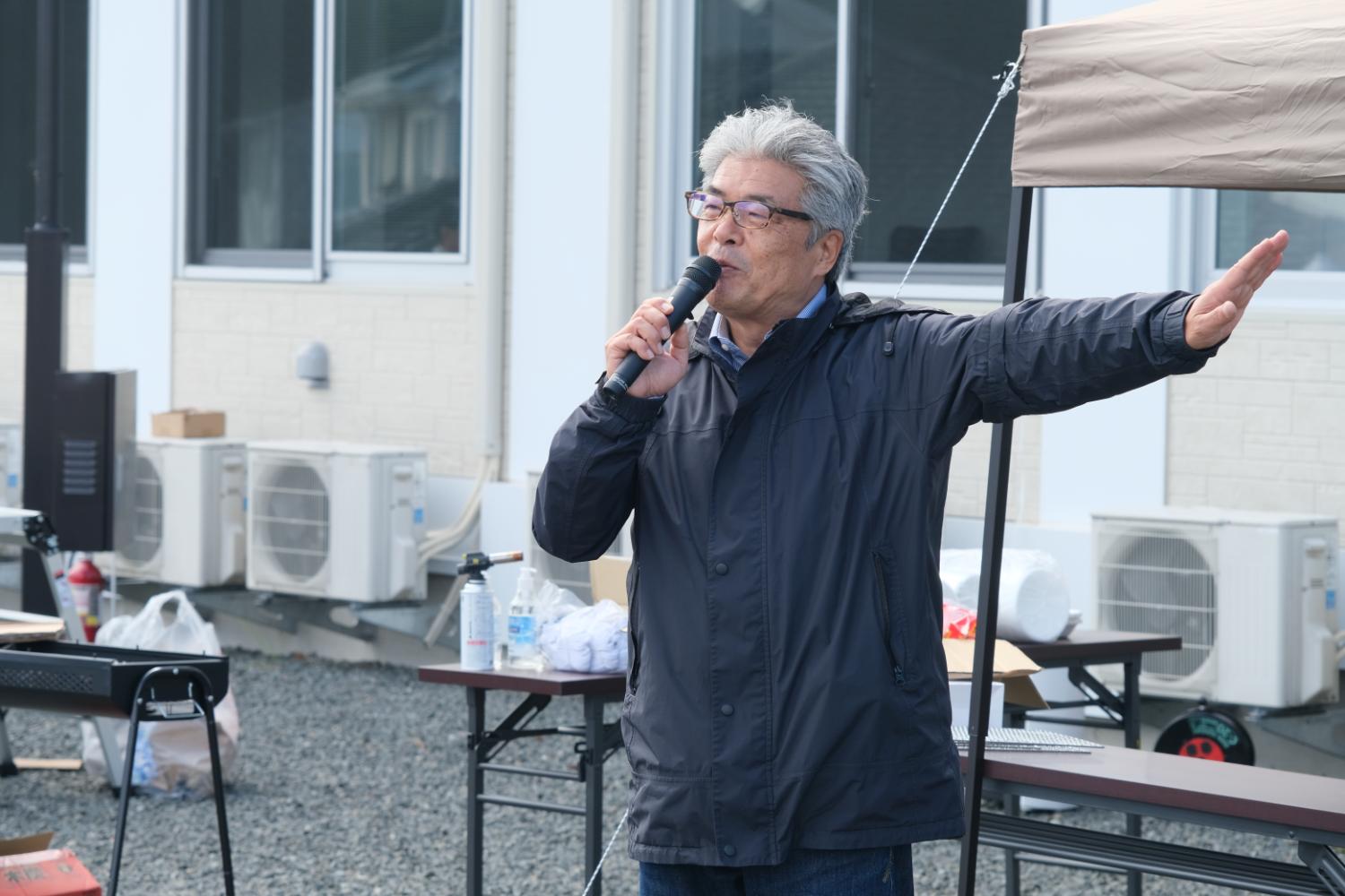 イベント会場の屋外で、山内さんがマイクを持ち、片腕を横に広げて参加者に語りかけている様子の写真