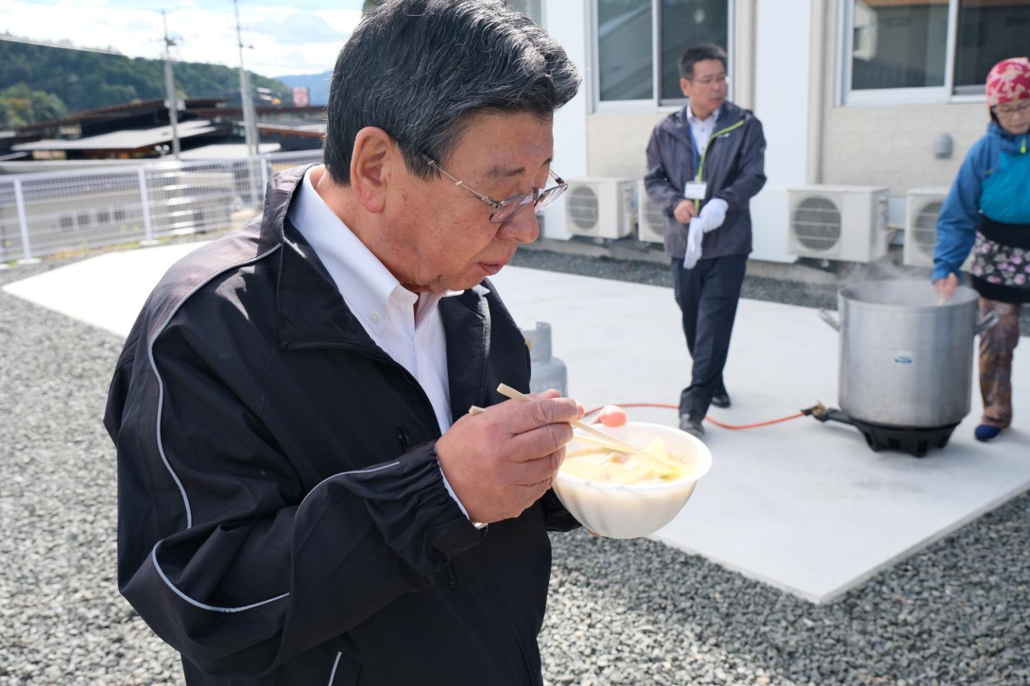 屋外で佐藤仁町長が黒いジャケットを着て白い皿を手に持ち、箸で温かい汁物を食べており、背景には調理用の大きな鍋と調理中の人がいる様子の写真